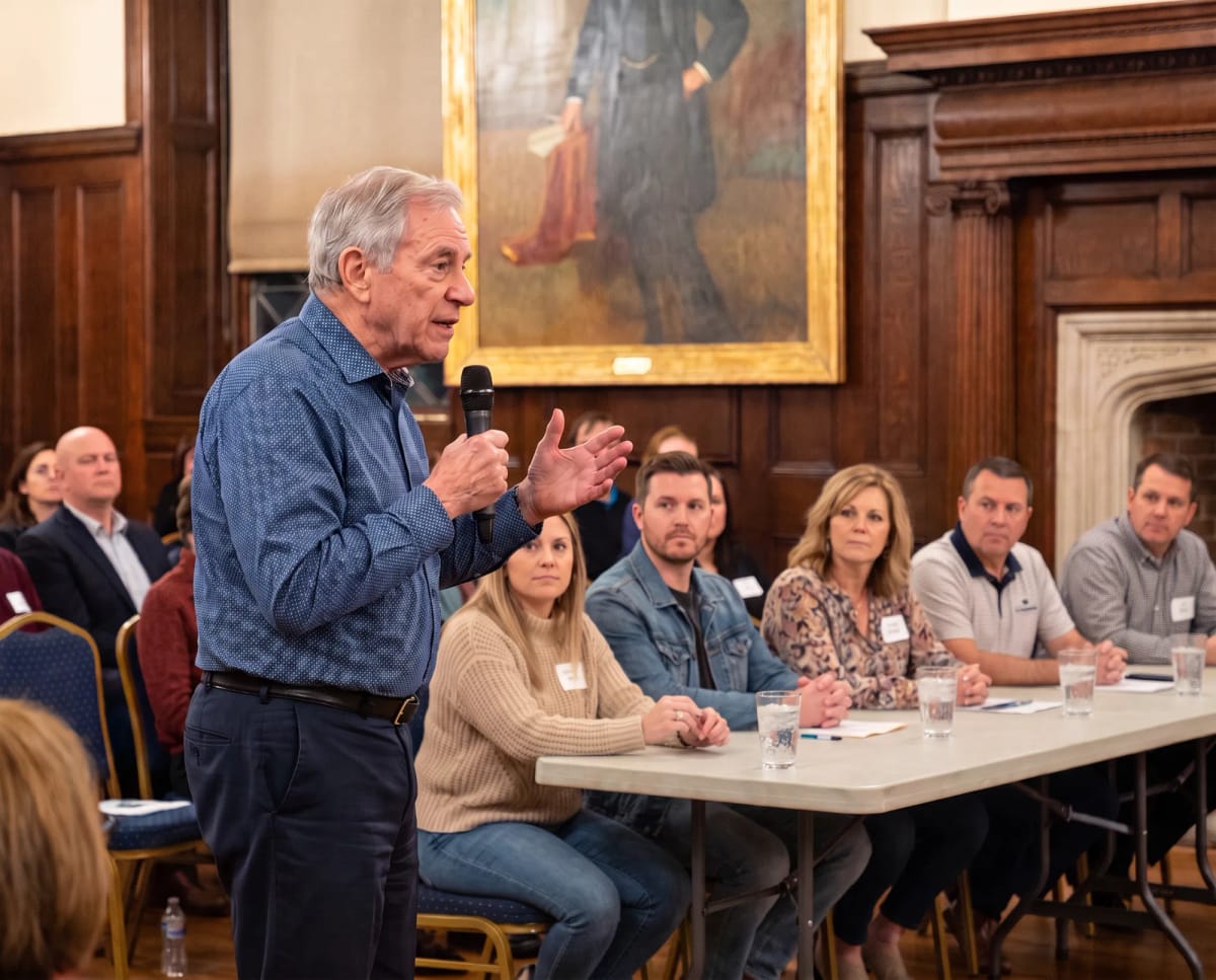 Man speaking into a microphone at a community meeting, with a panel seated at a table